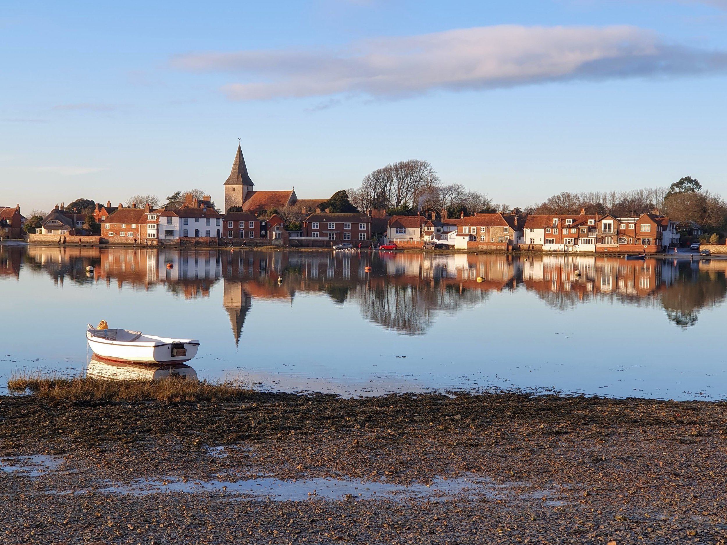 Bosham harbour at slack water: the church spire and waterfront cottages reflected in still water, a white rowing boat on the shingle